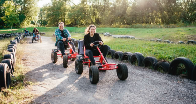 Two people riding red pedal go-karts on a dirt track lined with tires, surrounded by greenery and trees.