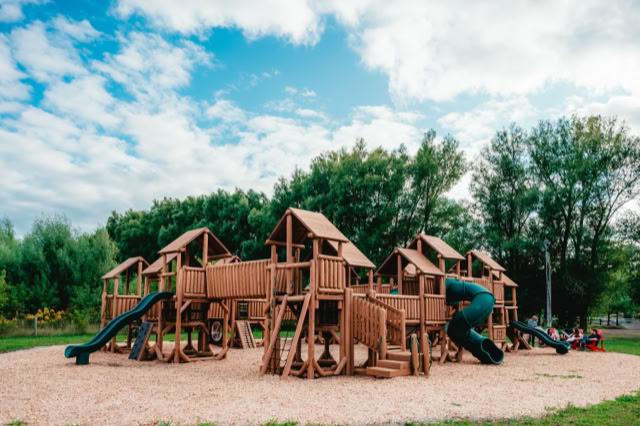 Wooden playground structure with slides and climbing areas, surrounded by trees and a grassy field under a partly cloudy sky.