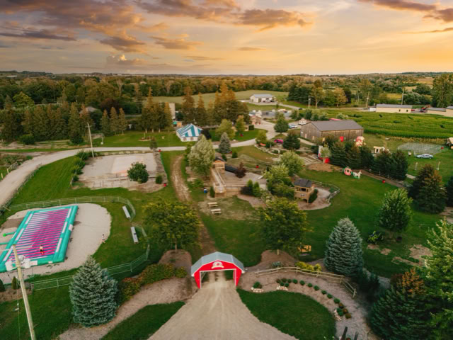 Aerial view of a rural landscape with a red barn, swimming pool, trees, and fields under a sunset sky.