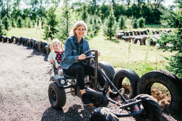 A woman and a child riding a pedal go-kart on a dirt path lined with tires, surrounded by greenery and trees.