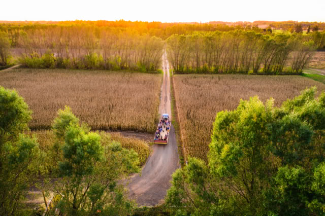 Aerial view of a rural road lined with trees, with a vehicle driving along it, surrounded by fields and a sunset in the background.
