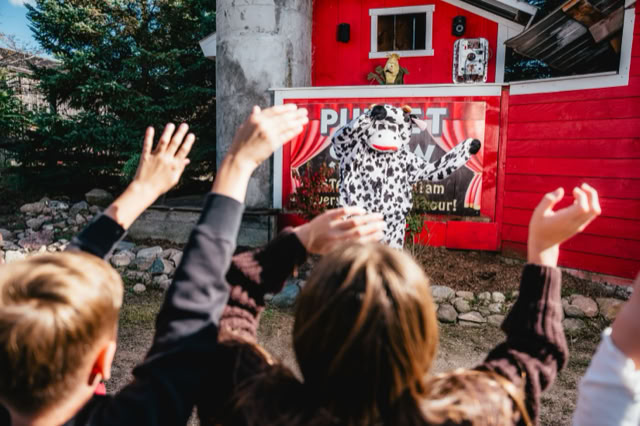 Children with raised hands facing a person in a cow costume standing in front of a red barn-like structure.
