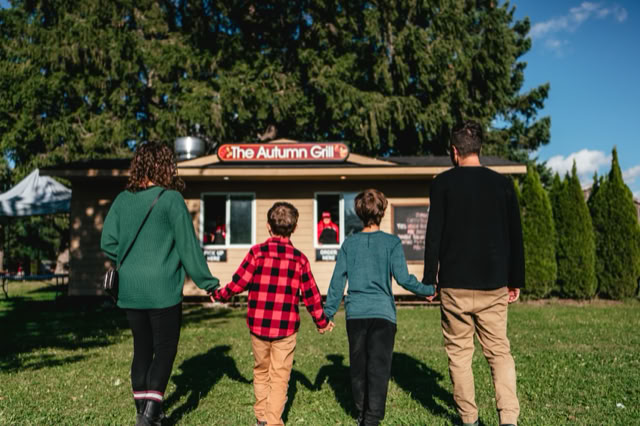 Four people holding hands, walking towards a small building with a sign that reads "The Autumn Grill," surrounded by trees and grass.