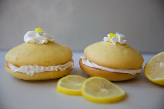 Two lemon-flavored whoopie pies with cream filling, topped with whipped cream and a small yellow candy, next to lemon slices.