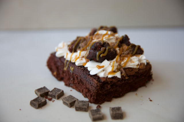 A piece of chocolate cake topped with whipped cream, chocolate chunks, and caramel drizzle, with additional chocolate pieces scattered on the surface.
